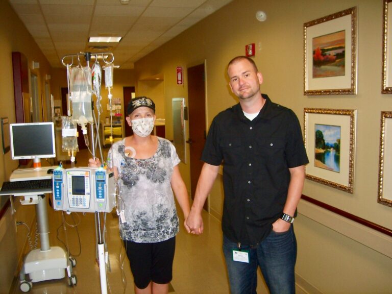 Two people holding hands in a hospital hallway beside IV equipment